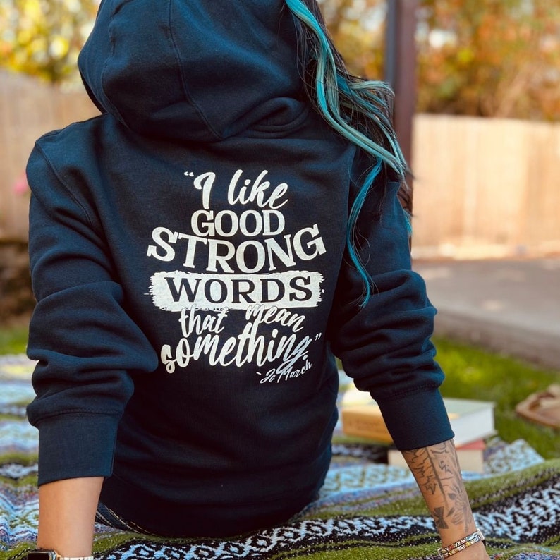 May include: A navy blue hoodie with a hood, featuring the quote "I like good strong words that mean something" in white lettering. The quote is attributed to Louisa May Alcott. The hoodie is worn outdoors.