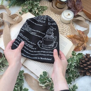May include: Black beanie featuring a white illustration of a teacup tower and text from "Alice's Adventures in Wonderland." The hat has printed text and is held over an open book. The beanie is a stylish accessory.