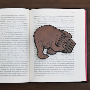 Puede incluir: Un marcapáginas con forma de oso pardo con la palabra "Winnie" visible, colocado en las páginas de un libro abierto. Un accesorio caprichoso para los amantes de los libros.
