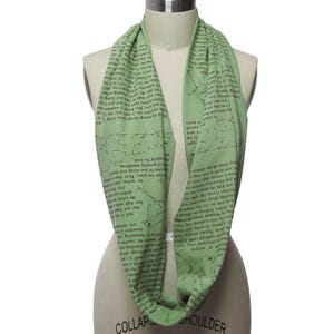 May include: A light green infinity scarf with a repeating pattern of text and bird illustrations. The scarf is wrapped around a mannequin, showcasing its circular design and soft texture. The text appears to be from a book.