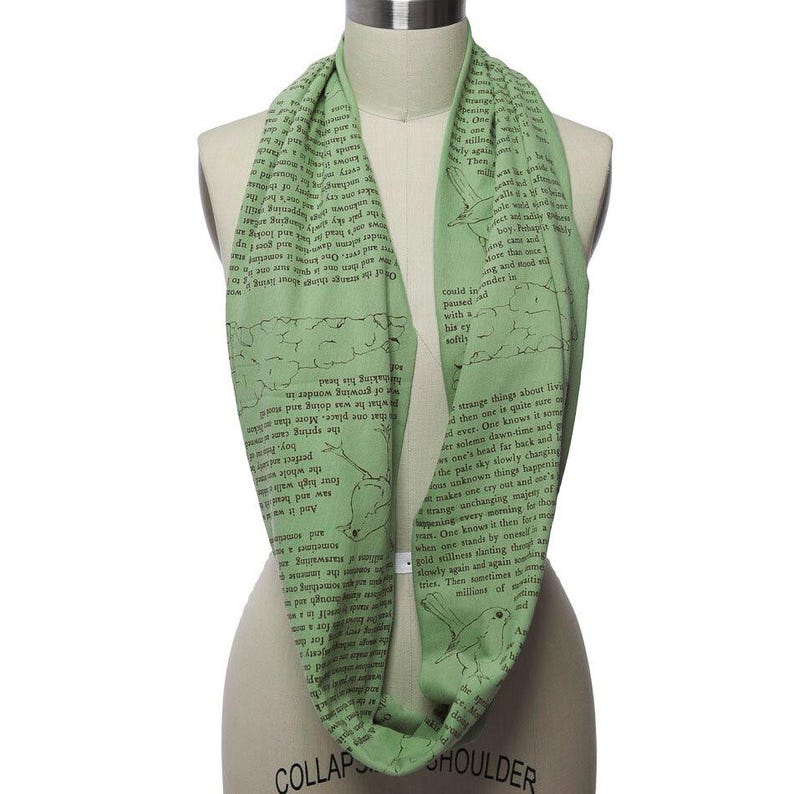May include: A light green infinity scarf with a repeating pattern of text and bird illustrations. The scarf is wrapped around a mannequin, showcasing its circular design and soft texture. The text appears to be from a book.