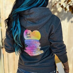 May include: Charcoal grey hoodie with a vibrant graphic on the back. The design shows a heart shape with the words "Book lover" and a stack of books in watercolour style. The hoodie has a drawstring hood.