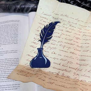 May include: A dark blue die-cut paper cutout of a quill pen resting in an inkwell, placed on top of a piece of aged, cream-colored parchment paper featuring handwritten cursive script and an open book with printed text in the background.