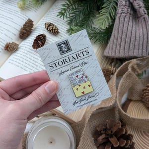 May include: A pin featuring a yellow envelope design with red heart accents and the words "Public Library" and "STORIARTS Superior Enamel Pins." The pin is held in front of a book, pinecones, and a knitted hat.