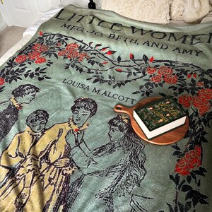 May include: A green throw blanket with a floral pattern and the title "Little Women" by Louisa M. Alcott. The blanket features a scene from the book with four girls.