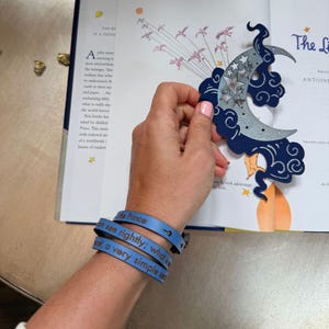 Puede incluir: Una pulsera de cuero azul con el texto "Little Prince" y una cita. Una luna decorativa y un recorte de nube con estrellas se sostienen sobre un libro abierto. El libro tiene ilustraciones y texto.
