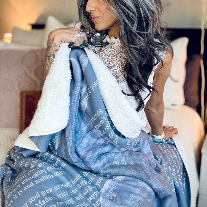 May include: A blue and white blanket with a soft, fluffy white underside. The blanket features a printed design with white text and a watercolor-like pattern. The blanket is draped over a bed.