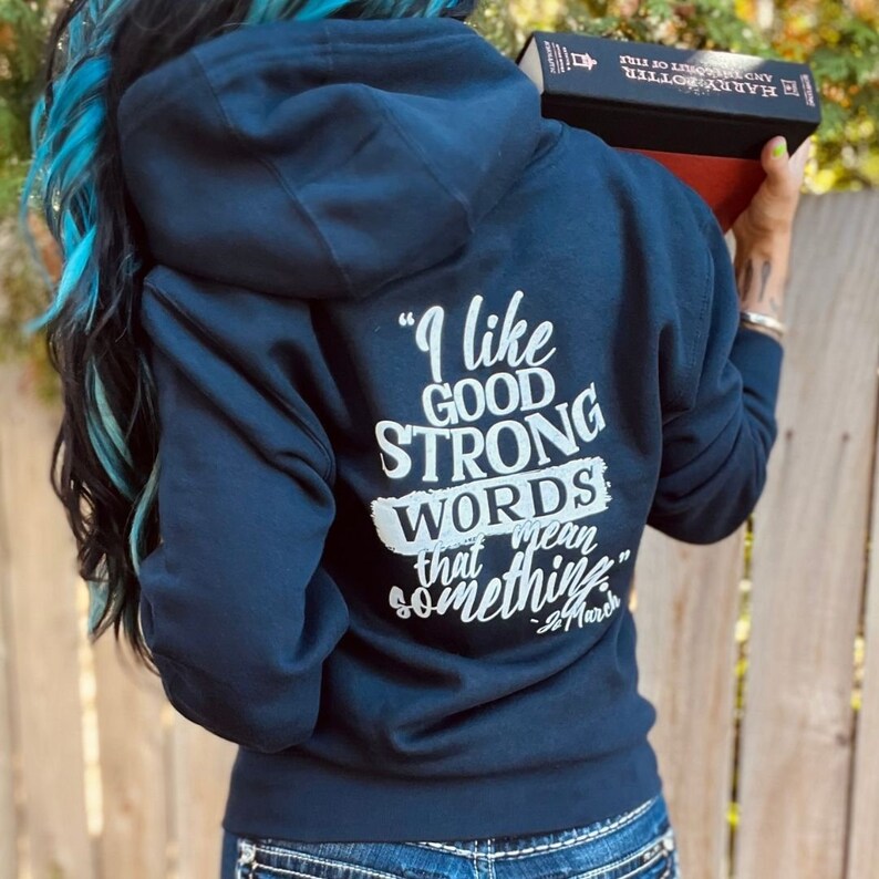 May include: Navy blue hoodie with a quote on the back that reads, "I like good strong words that mean something." The quote is in white lettering. The hoodie has a hood and is worn by a person with blue highlights in their hair.