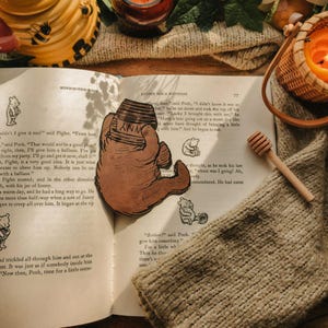 Peut inclure: Une découpe en cuir brun représentant un ours tenant un pot de miel avec l'inscription HUNNY, posée sur une page ouverte d'un livre Winnie l'ourson. Une cuillère à miel en bois repose sur la page à côté de la découpe aux détails gravés.