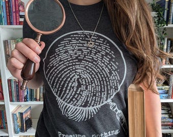 May include: A dark gray t-shirt with a magnifying glass design and the words "Presume nothing." A copper and wood magnifying glass is held up, and a book is held in the other hand. Bookshelves are in the background.
