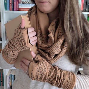 May include: Tan infinity scarf and matching fingerless gloves, both adorned with a script-like text pattern in black. The gloves have black trim. A book is held in the center. The scarf is wrapped around the neck.