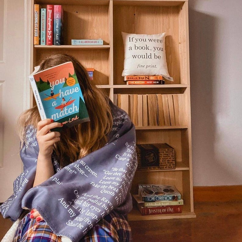May include: A person holding a book titled "You Have a Match" in front of their face, wrapped in a blue blanket with white text. A wooden bookshelf filled with books and a decorative pillow with text is in the background.