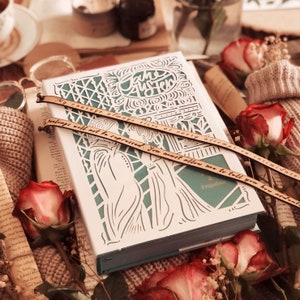 Peut inclure: Un livre décoratif avec une couverture blanche et un design découpé complexe, avec le titre "Pride & Prejudice". Deux marque-pages en bois avec des citations sont placés dessus. Des roses rouges et un article tricoté entourent le livre.