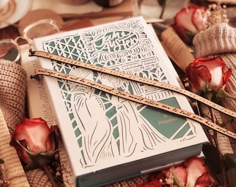 May include: A decorative book with a white cover and intricate cut-out design, featuring the title "Pride & Prejudice." Two wooden bookmarks with quotes are placed on top. Red roses and a knitted item surround the book.