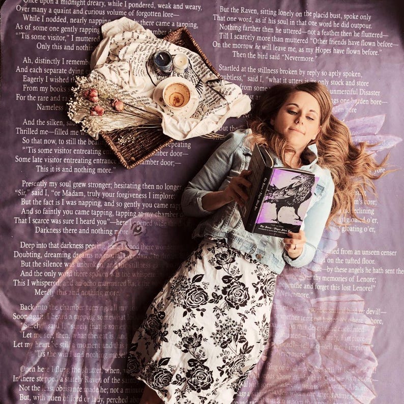 May include: A woman reads a book with a black cover while lying on a purple patterned surface. A woven tray with dried flowers, a cup, and candles is beside her. She wears a denim jacket and a black and white floral skirt.
