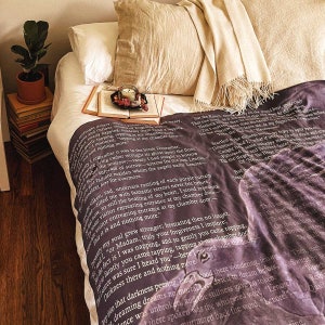 May include: A bed with white sheets, a beige pillow, and a patterned blanket with text. A small potted plant, books, and a candle on a tray are placed nearby. The blanket has a dark purple background.