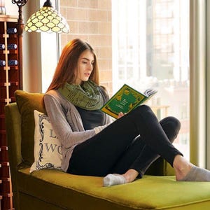 May include: A woman reading a book while sitting on a green chaise lounge. She is wearing a gray cardigan, black pants, and a green scarf. A decorative pillow with text is behind her. A stained glass lamp hangs above.