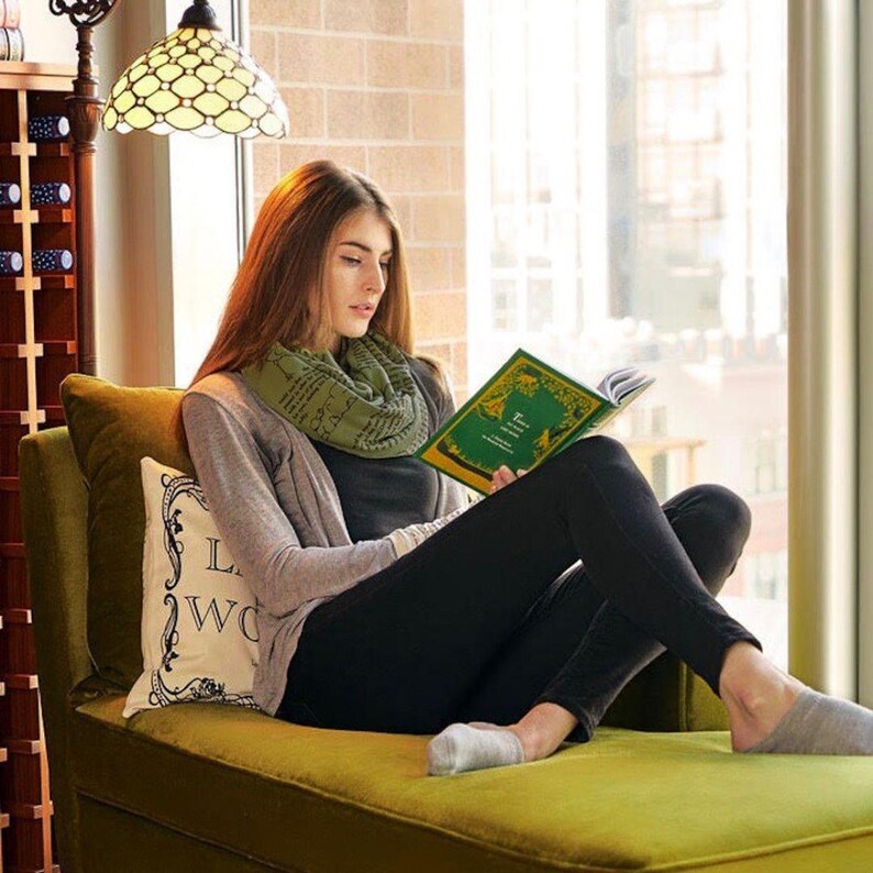 May include: A woman reading a book while sitting on a green chaise lounge. She is wearing a gray cardigan, black pants, and a green scarf. A decorative pillow with text is behind her. A stained glass lamp hangs above.