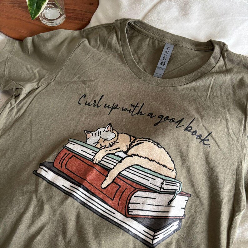 Puede incluir: Camiseta verde oliva con una ilustraci&oacute;n de un gato durmiendo sobre una pila de libros. El texto "Curl up with a good book" est&aacute; impreso encima del gato. La camiseta es de un material suave.