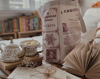 May include: A canvas tote bag with the text "A Christmas Carol" by Dickens, a teapot, teacup, and open book. The tote bag has a snowflake design and burgundy text. The teacup and teapot are white with gold accents.