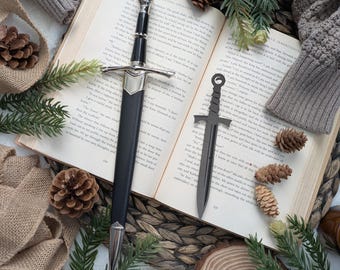 Puede incluir: Dos espadas decorativas plateadas y negras, una con mango negro y la otra con mango plateado, se colocan sobre un libro abierto con un fondo de mimbre marrón. El libro está abierto en una página con texto y una estera tejida marrón y blanca es visible en el fondo.