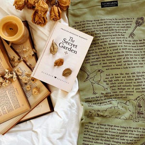 May include: A still-life arrangement featuring a copy of "The Secret Garden" book, dried leaves, and a lit candle. A green tote bag with text and illustrations is also present, creating a vintage aesthetic.