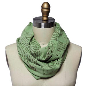 May include: A light green infinity scarf with a printed design featuring text and illustrations. The scarf is draped on a mannequin, showcasing its circular shape and soft fabric. The design includes a key and a door.