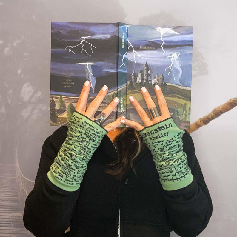 Puede incluir: Un libro de tapa dura titulado "Frankenstein" de Mary Shelley, con una ilustraci&oacute;n dram&aacute;tica de un castillo y rel&aacute;mpagos. La imagen tambi&eacute;n presenta calentadores de brazo verdes con texto y una sudadera con capucha negra.