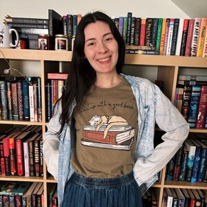 Puede incluir: Una mujer con una camiseta marr&oacute;n con el texto "Curl up with a good book" y un dibujo de un gato durmiendo sobre libros. Est&aacute; de pie frente a una estanter&iacute;a llena de libros. Tambi&eacute;n lleva una chaqueta vaquera y una falda vaquera.