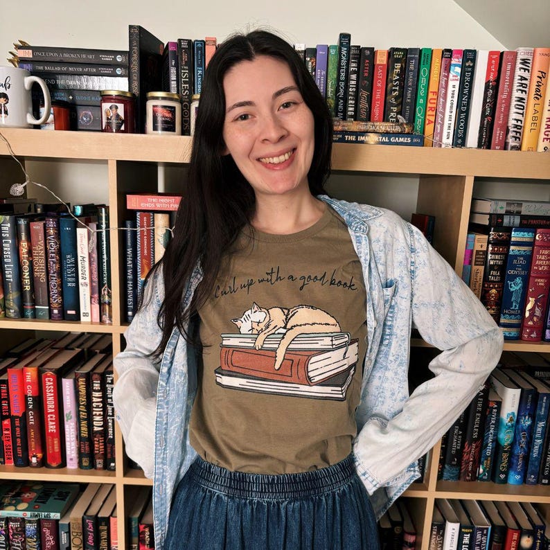Puede incluir: Una mujer con una camiseta marr&oacute;n con el texto "Curl up with a good book" y un dibujo de un gato durmiendo sobre libros. Est&aacute; de pie frente a una estanter&iacute;a llena de libros. Tambi&eacute;n lleva una chaqueta vaquera y una falda vaquera.