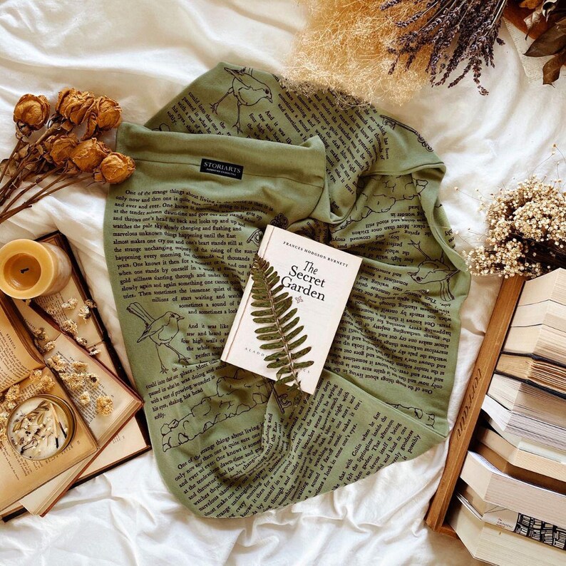 May include: An olive green long-sleeve shirt with a bird illustration and text from a book. The book "The Secret Garden" is open, with a fern and dried flowers nearby. The shirt has the brand name "STORIARTS" on it.