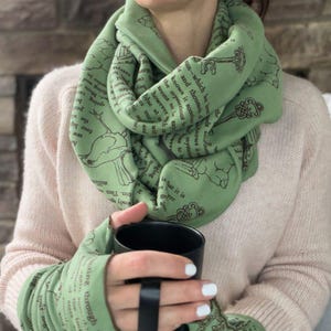 May include: A green infinity scarf and matching fingerless gloves. The scarf and gloves feature a repeating pattern of keys, birds, and text. The person is holding a black mug. The scarf is wrapped around the neck.
