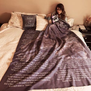 May include: A dark grey throw blanket with text from a poem, draped over a bed. The blanket features a detailed illustration and the words "The Raven" in white. A decorative pillow with "Sense & Sensibility" is also on the bed.