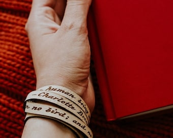 May include: A beige leather bracelet with the quote "Human being - Charlotte Bronte - no bird; and no" engraved on it.