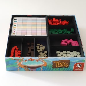 May include: A board game box organizer for Tiny Towns containing a grid score sheet and various colored wooden resource tokens including red, green, pink, grey, brown, and tan shapes separated into black compartments within the cardboard box.