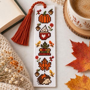 Puede incluir: Un marcapáginas rectangular blanco con un patrón de punto de cruz de motivos otoñales que incluye una calabaza naranja, una taza roja con un corazón blanco, una pila de libros, una hoja de arce y un erizo, acabado con una borla de hilo color óxido.