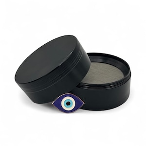 Puede incluir: Un molinillo de hierbas cilíndrico negro con agarre texturizado y una malla metálica fina en el interior. Frente al molinillo hay un pequeño pin de esmalte con forma de ojo que presenta círculos concéntricos en azul oscuro, blanco y azul claro.