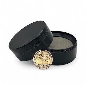 May include: A black cylindrical herb grinder with a textured grip edge, shown with its lid removed to reveal a fine metal mesh screen. A circular bronze-toned medallion featuring a raised relief of a mountain range and forest sits in front.