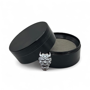 May include: A black cylindrical herb grinder with a textured grip edge and a fine metal mesh screen inside. A silver-toned, detailed Hannya mask emblem with prominent horns and sharp teeth is attached to the front of the grinder's lid.