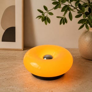 Peut inclure: Une lampe de table circulaire en forme de beignet avec un corps translucide orange lumineux. Le centre de la lampe présente un accent métallique argenté lisse et réfléchissant. La lampe repose sur une base circulaire noire solide.