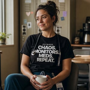 Puede incluir: Una camiseta negra de manga corta con el texto blanco ICU NURSE CHAOS. MONITORS. MEDS. REPEAT. La prenda se lleva con un estetoscopio plateado alrededor del cuello y pantalones de trabajo azul oscuro, mientras se sostiene una taza blanca.