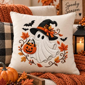May include: A square off-white throw pillow featuring an embroidered design of a white ghost wearing a black witch hat with an orange bow. The ghost holds an orange jack-o'-lantern bucket filled with candy, surrounded by orange maple leaves and bats.