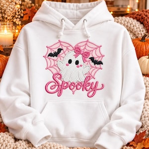 May include: A white hooded sweatshirt featuring an embroidered design of a white ghost with a pink bow, centered within a pink spiderweb heart. Two black bats flank the ghost, and the word Spooky is embroidered in pink script below the heart.