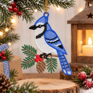 May include: A blue jay bird ornament featuring detailed embroidery in shades of blue, white, and black. The bird is perched on a brown branch with green pine needles, red berries, and a small brown pinecone, suspended by a gold string.