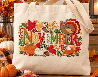 May include: A cream-colored canvas tote bag featuring an embroidered design with the word NOVEMBER in colorful letters, surrounded by autumn motifs including a turkey, pumpkins, fall leaves, acorns, wheat stalks, and a slice of pumpkin pie.