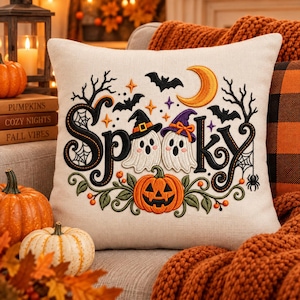 May include: A square throw pillow with a cream-coloured fabric cover featuring an embroidered design. The text Spooky is written in black script, surrounded by two ghosts wearing hats, a jack-o'-lantern, bats, a crescent moon, and spiderwebs.