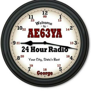 May include: A round wall clock with a black frame and a cream-colored face. The dial features 12-hour and 24-hour markers, black hands, and the text Welcome to AE63YA 24 Hour Radio Your City, State's Best George in black and red lettering.