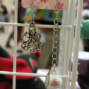 Puede incluir: Un par de pendientes de plata con un pequeño encanto de pulpo en uno y un pequeño encanto de taza de té en el otro. Los pendientes cuelgan de una tarjeta blanca con un patrón floral rosa.