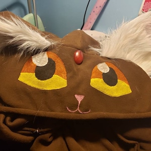 May include: Brown hooded sweatshirt with a cat face design. The cat has white fur, yellow eyes, and a pink nose.