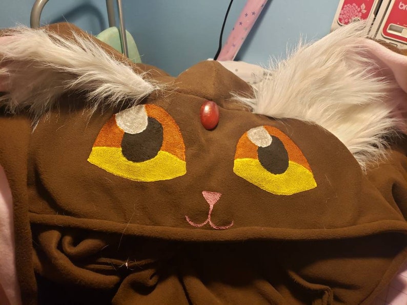 May include: Brown hooded sweatshirt with a cat face design. The cat has white fur, yellow eyes, and a pink nose.